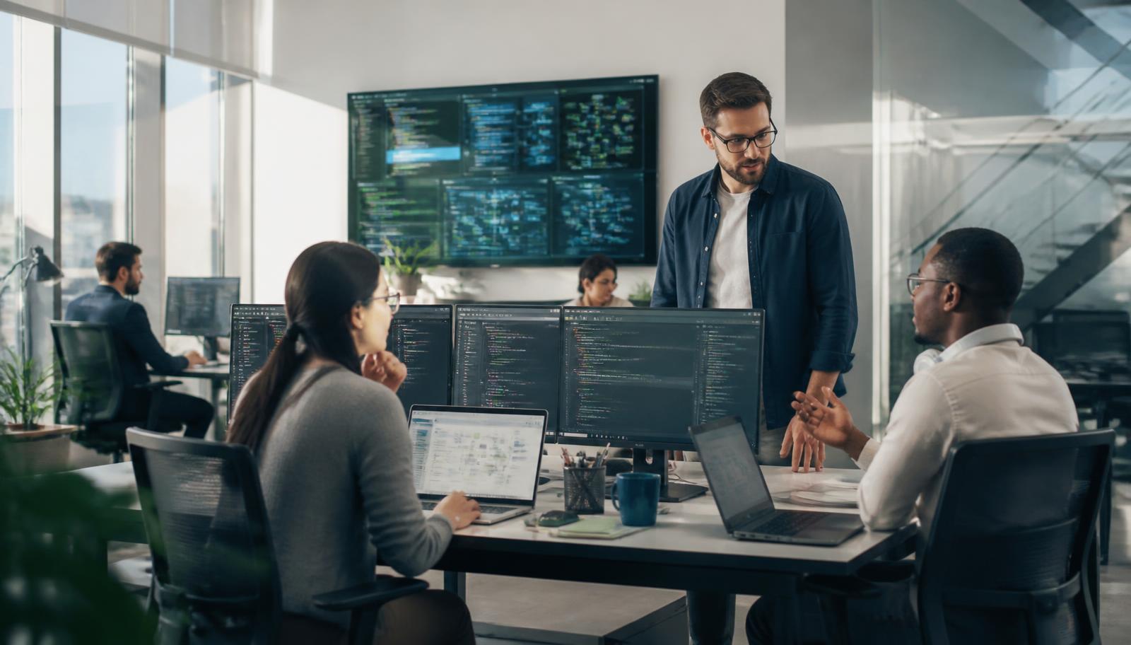 Software team collaborating around monitors showing code and dashboards in a bright modern office.